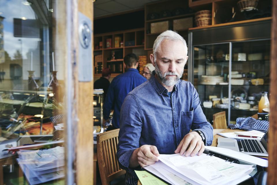 Mature sales clerk reading documents while sitting at table in deli