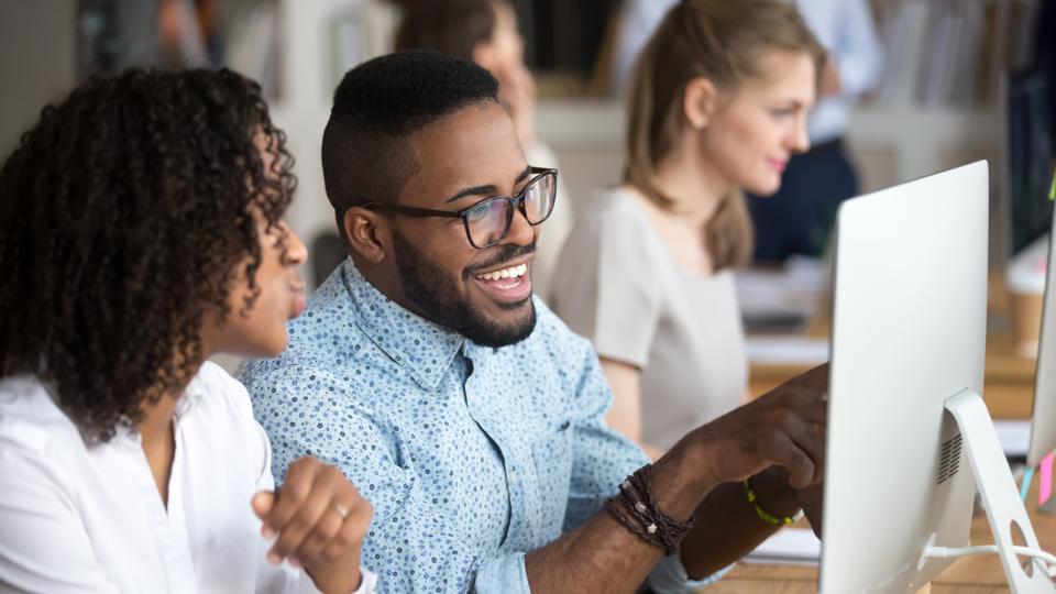 Smiling happy African American man showing funny news to female colleague, looking at screen, employee laughing on workplace, using computer, excited by good result of work