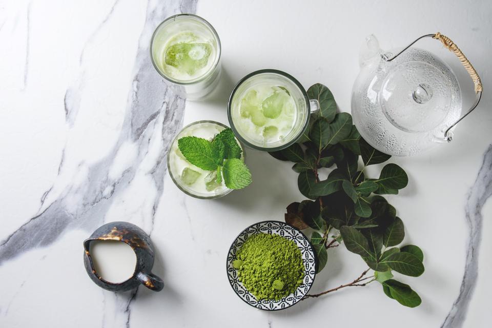 Matcha green tea iced latte or cocktail in three different glasses with ice cubes, matcha powder and jug of milk on white marble table, decorated by green branches Grey wall at background