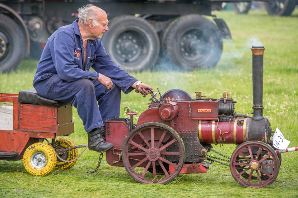 Mature male in blue jumpsuit rides along on miniature tractor steam engine on grass.
