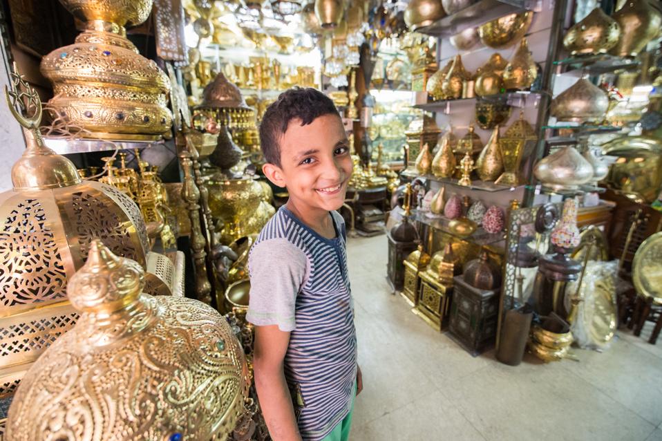 Young Egyptian boy standing in a shop selling ornate gold pots and vases in the outdoor bazaar.