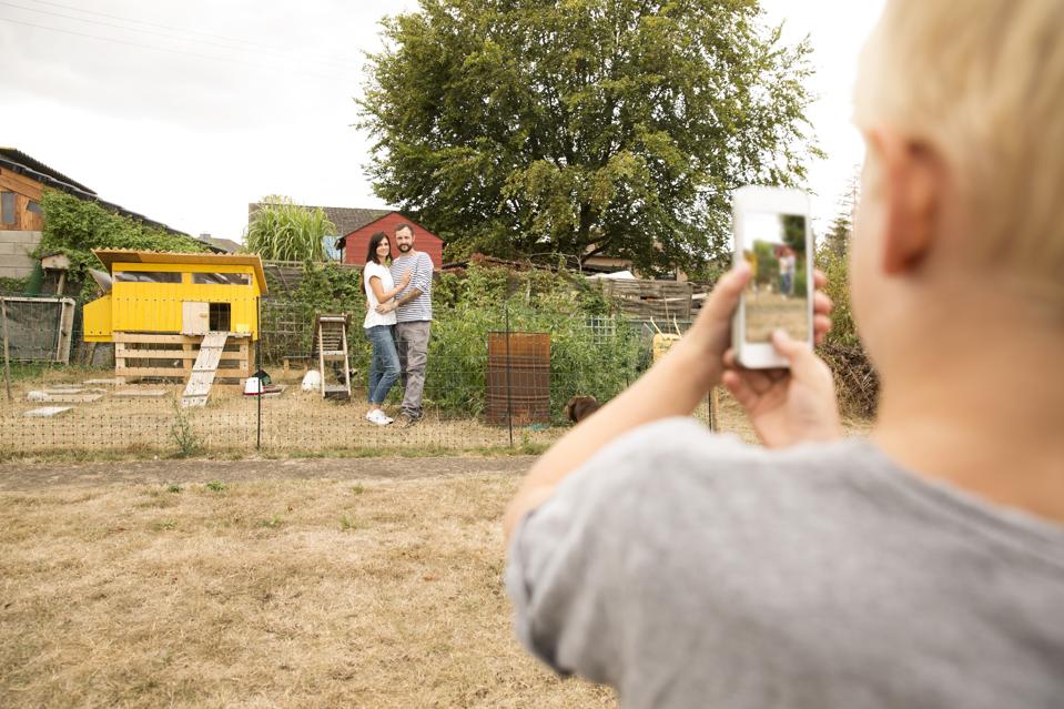 Boy taking cell phone picture of parents standing at chickenhouse in garden