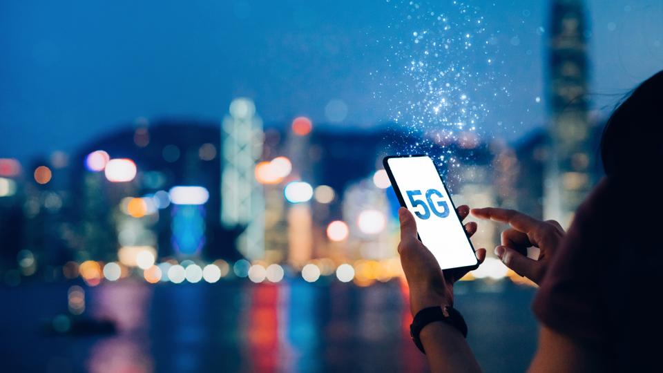 Young woman using smartphone against the iconic city skyline of Hong Kong by the promenade of Victoria Harbour at night, with the concept of 5G communications dissolved into light particles