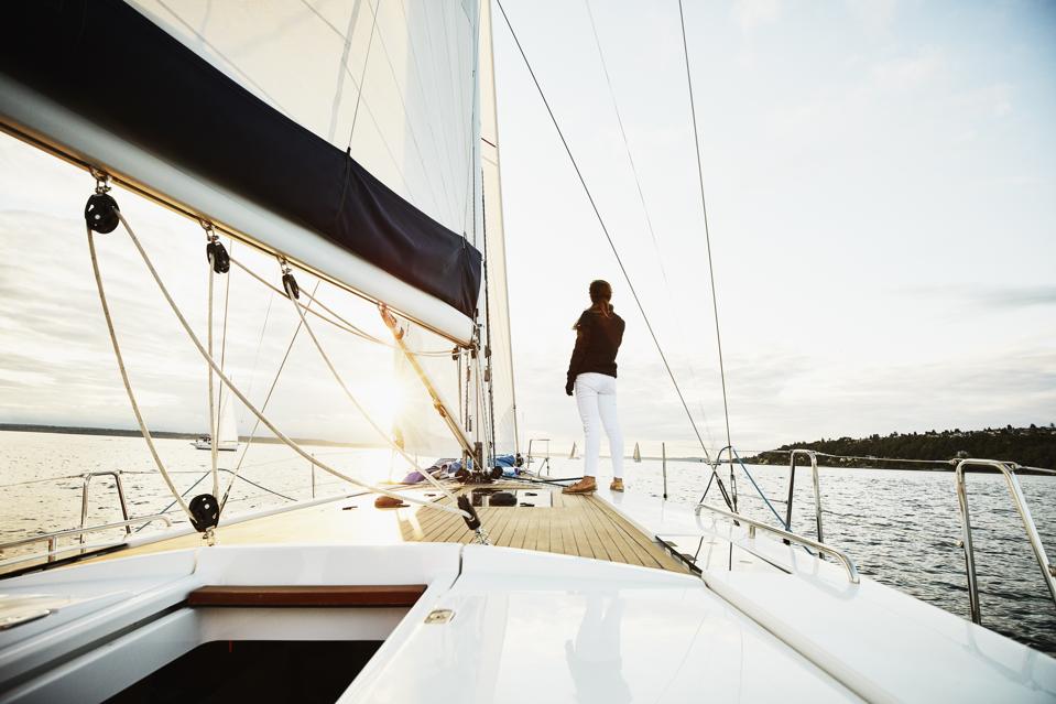 Female sailor standing on foredeck of sailboat watching sunset on summer evening
