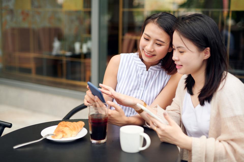 Two young women having coffee break together use smart phone. Happy women using cell phone at sidewalk cafe.