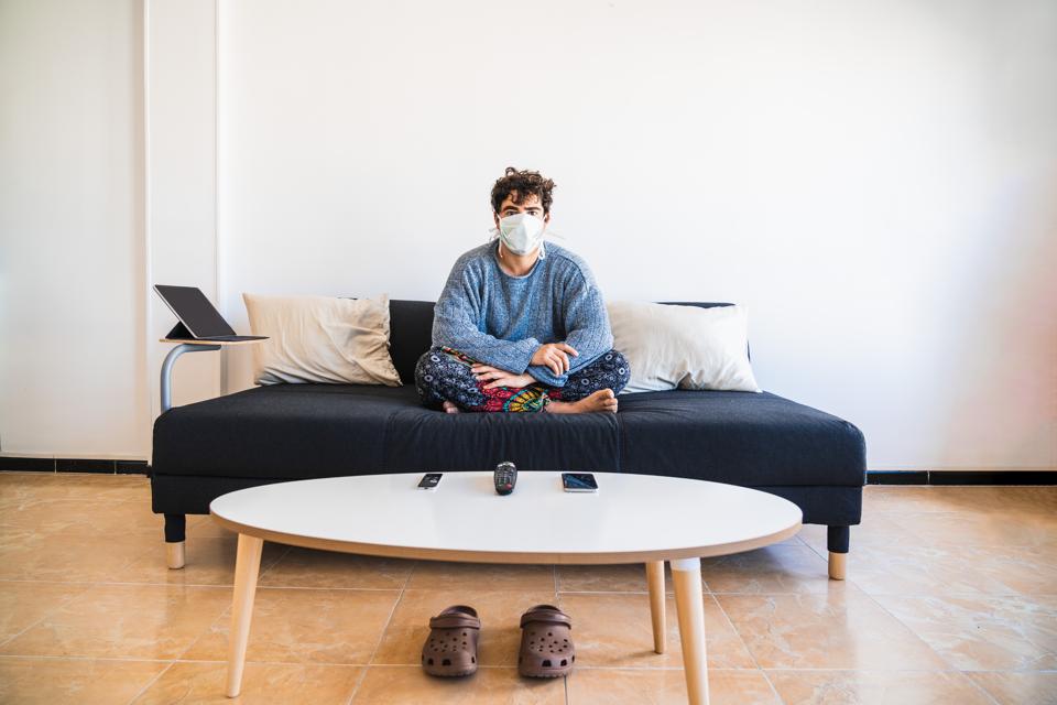 Young hispanic man sits on the couch while protecting against COVID-19 coronavirus due to it’s virulency and impact on media and society