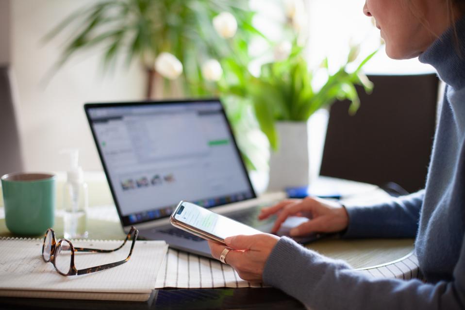 Woman working from home using laptop computer while reading text message on mobile phone