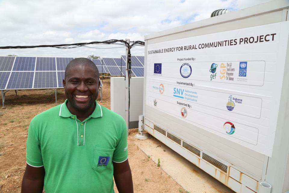 A man in a green shirt smiles in front of solar panels