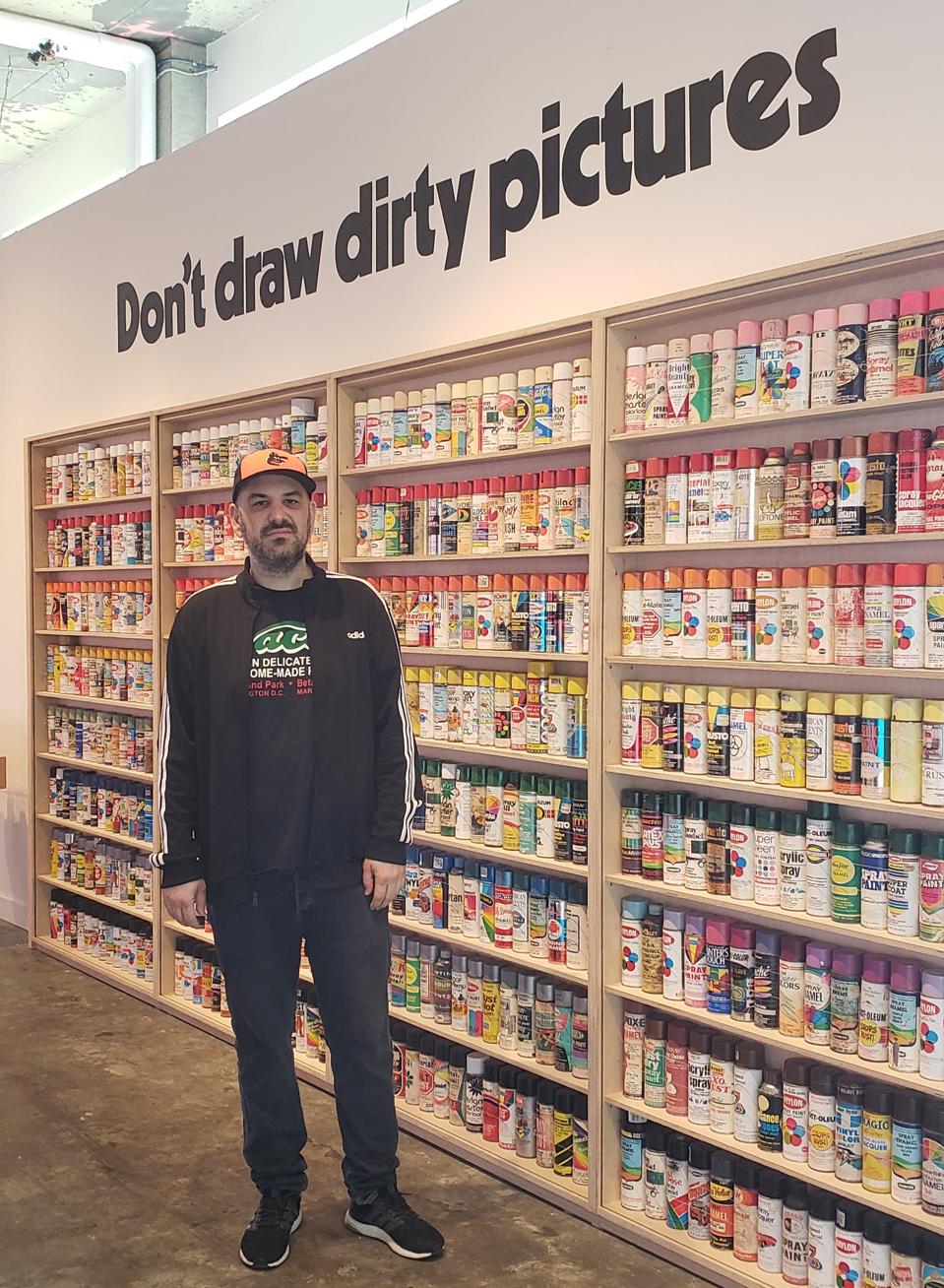 Roger Gastman in front of old spray paint cans; the wall features an ad campaign slogan from one of those brands of spray paint in the 1980s.