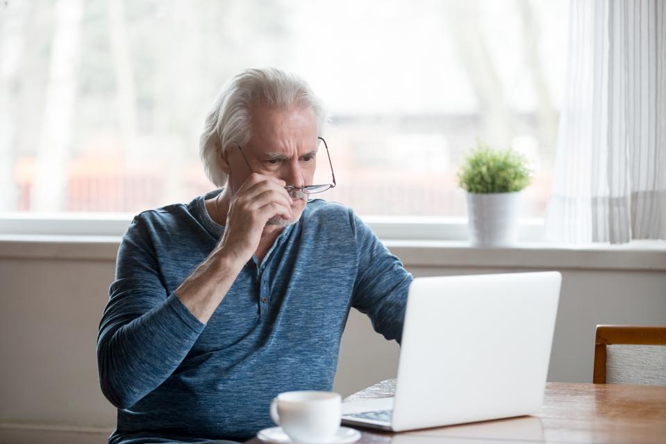 Shocked frustrated senior man taking off glasses looking at laptop