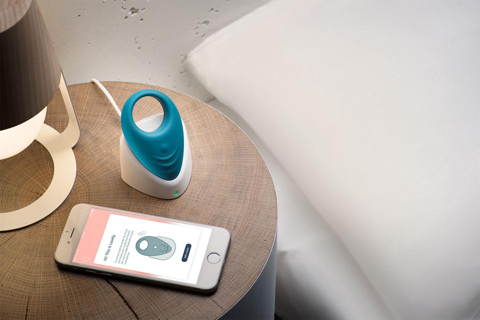 Lovely 2.0 wearable sex tech charing on a bedside table next to a smartphone