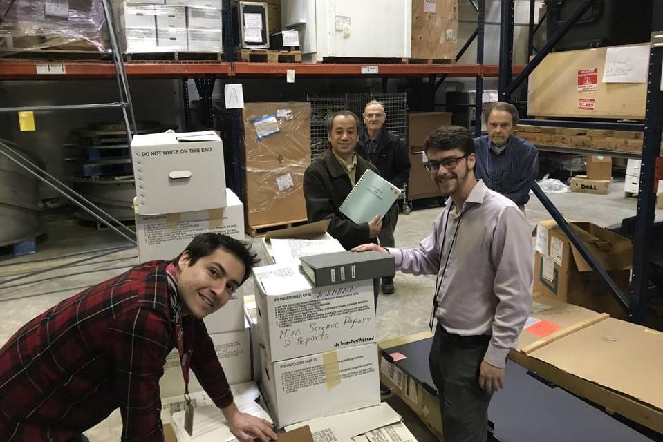 Color photo of a group of male engineers and scientists standing in a warehouse with a stack of boxes.