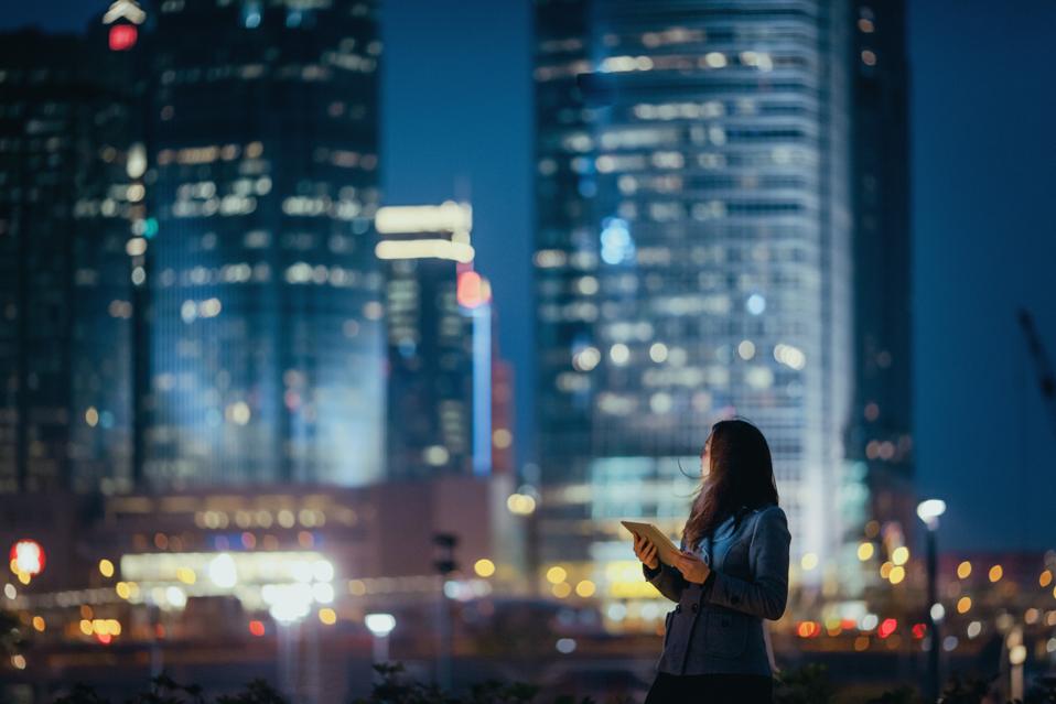 Young businesswoman using digital tablet, looking back over illuminated city skyline with confidence at night
