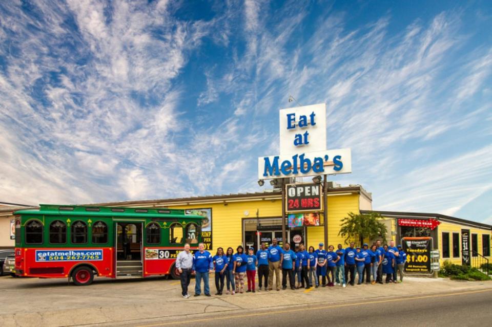 A restaurant with a bus that looks like a trolly and about twenty employees standing out front below a sign that reads ″Eat at Melba's.″
