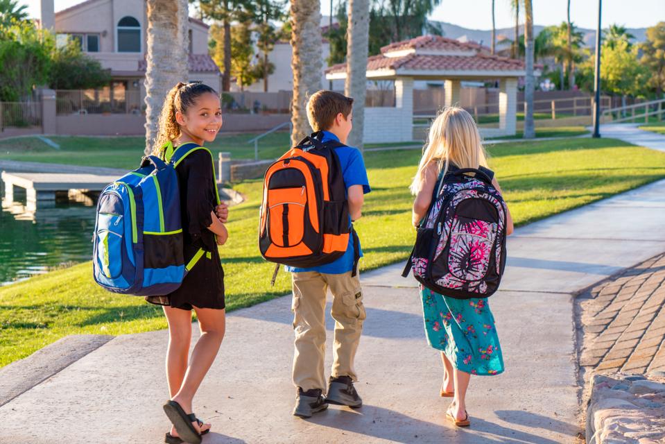Three grade school children wearing colorful backpacks walk together along a path on a sunny morning.