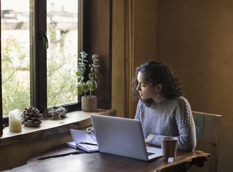 Young Woman of Mixed-ethnicity Works From Home Using Laptop Computer and Reference Book