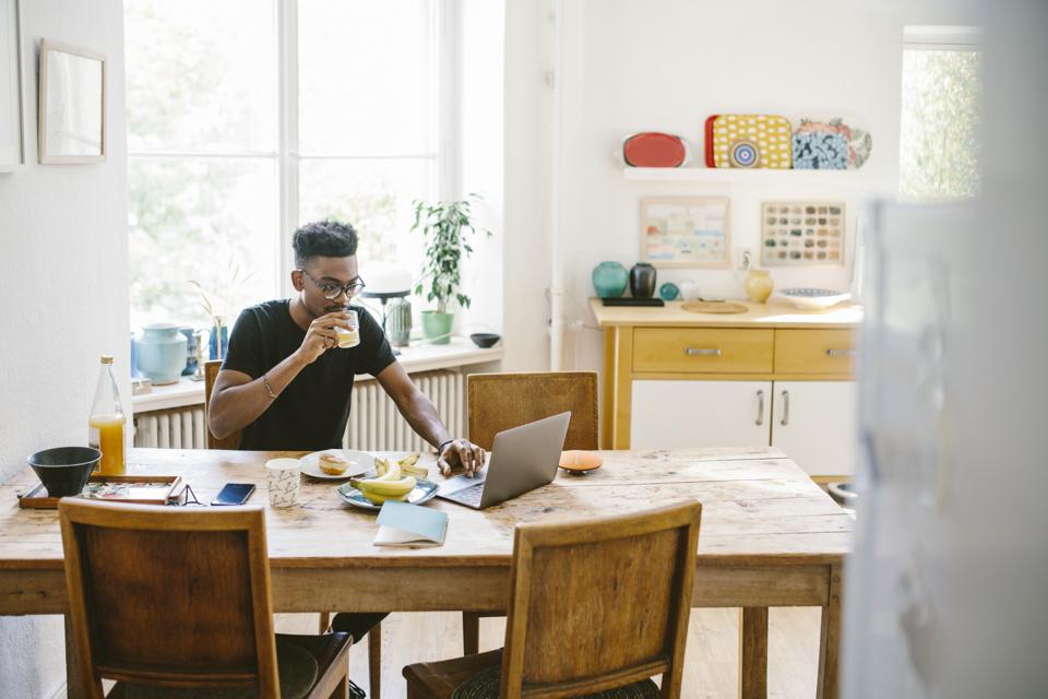 Young man drinking juice while using laptop at table in house