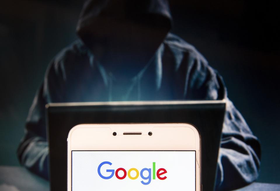Hooded hacker sitting at laptop with smartphone displaying the Google logo in foreground