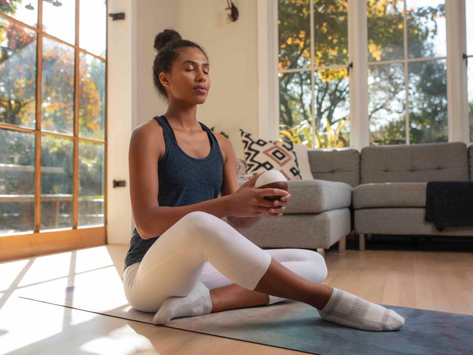 A young woman sits cross-legged meditating on a yoga mat with a white egg-like device