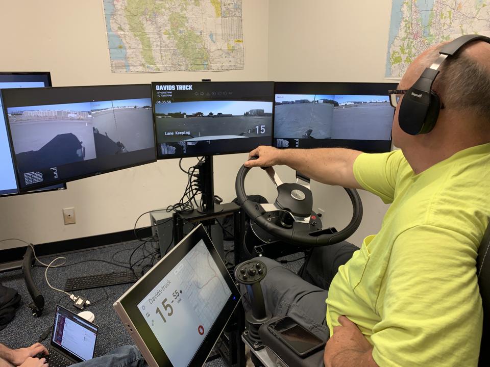 A truck driver sits behind a series of screens and remotely drives a truck