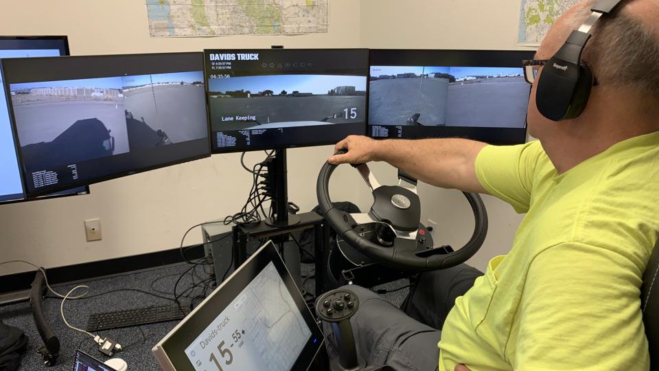 A truck driver sits behind a series of screens and remotely drives a truck