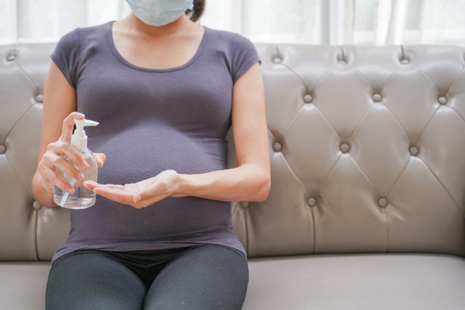 A pregnant woman wears a surgical mask is washing hands by alcohol gel on the sofa. Protect a COVID-19 (Coronavirus)