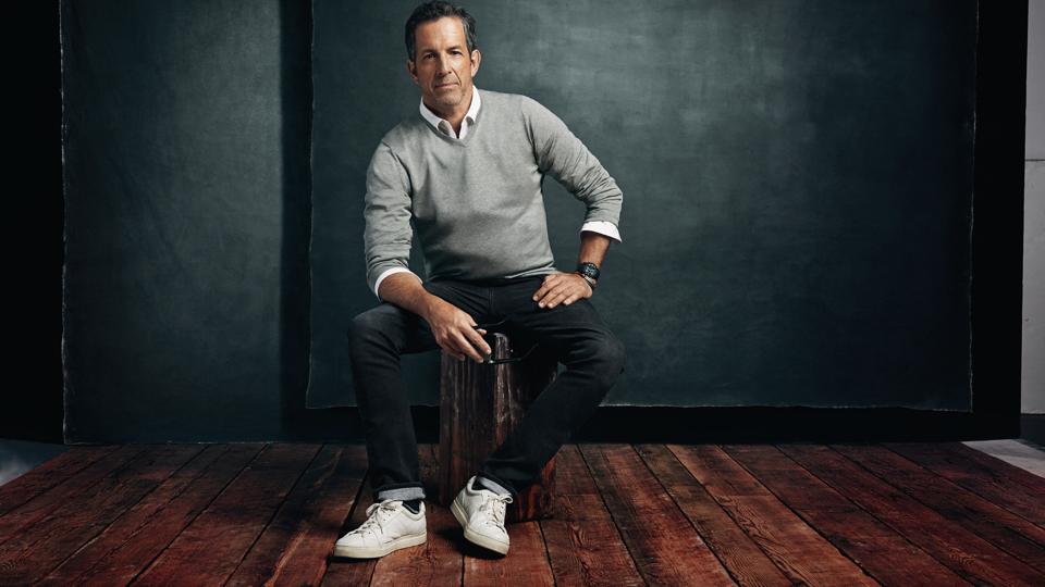 Fashion designer Kenneth Cole sitting on a wooden stool in a studio.