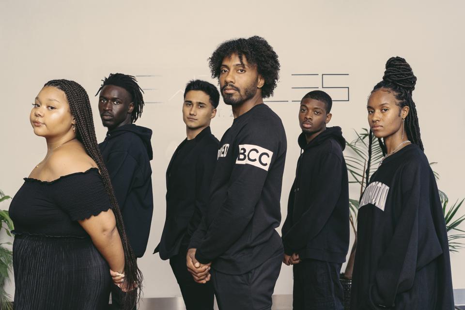Six young people, all dressed in black, stand with shoulders turned but faces looking at the camera.
