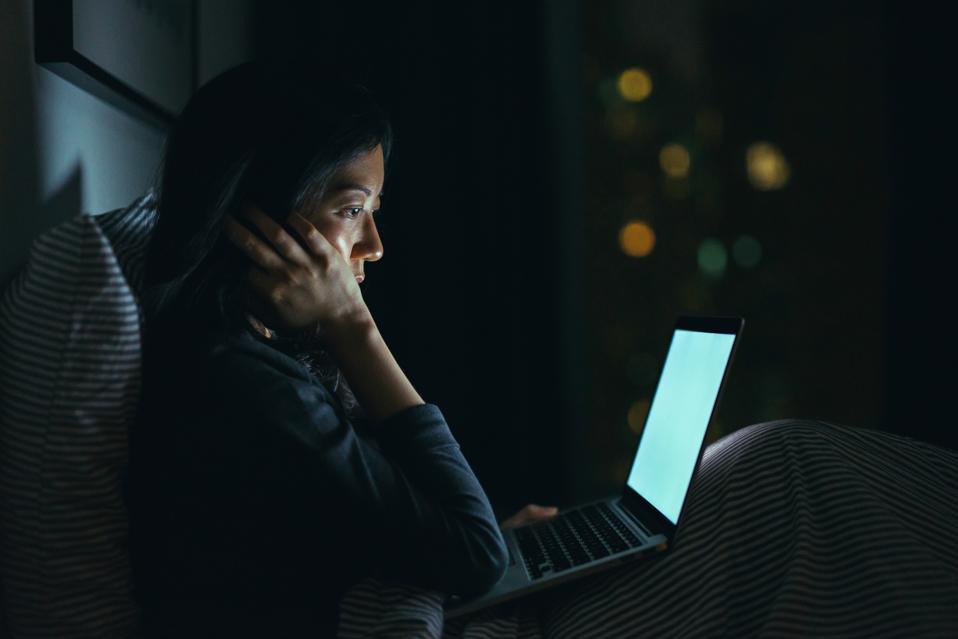 Young woman in deep thought while using laptop on bed at night
