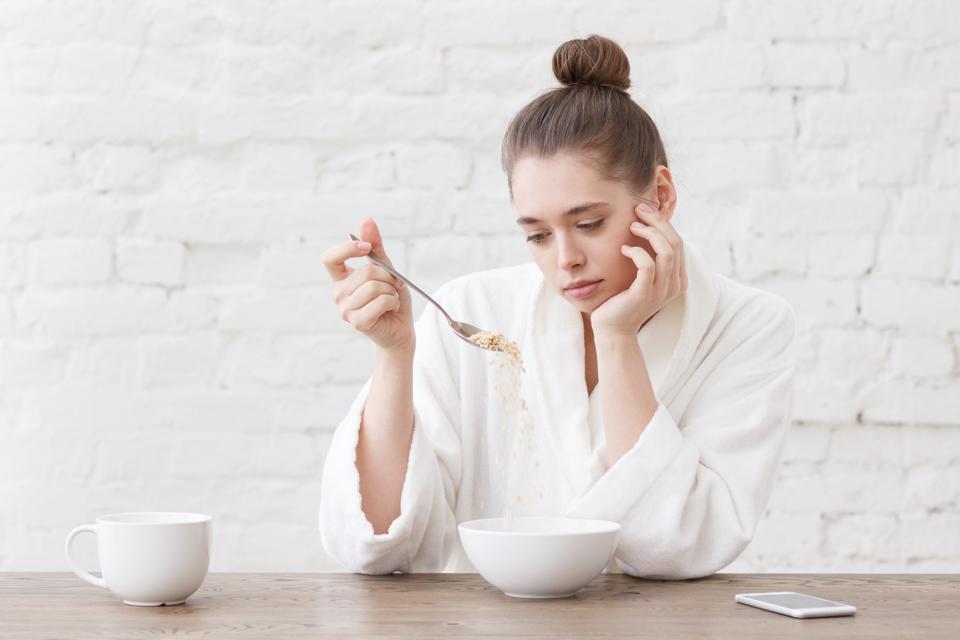 Young woman with no appetite, tired, bad mood and sleepy, sitting in the white loft kitchen, having unsavory unpalatable unappetizing breakfast