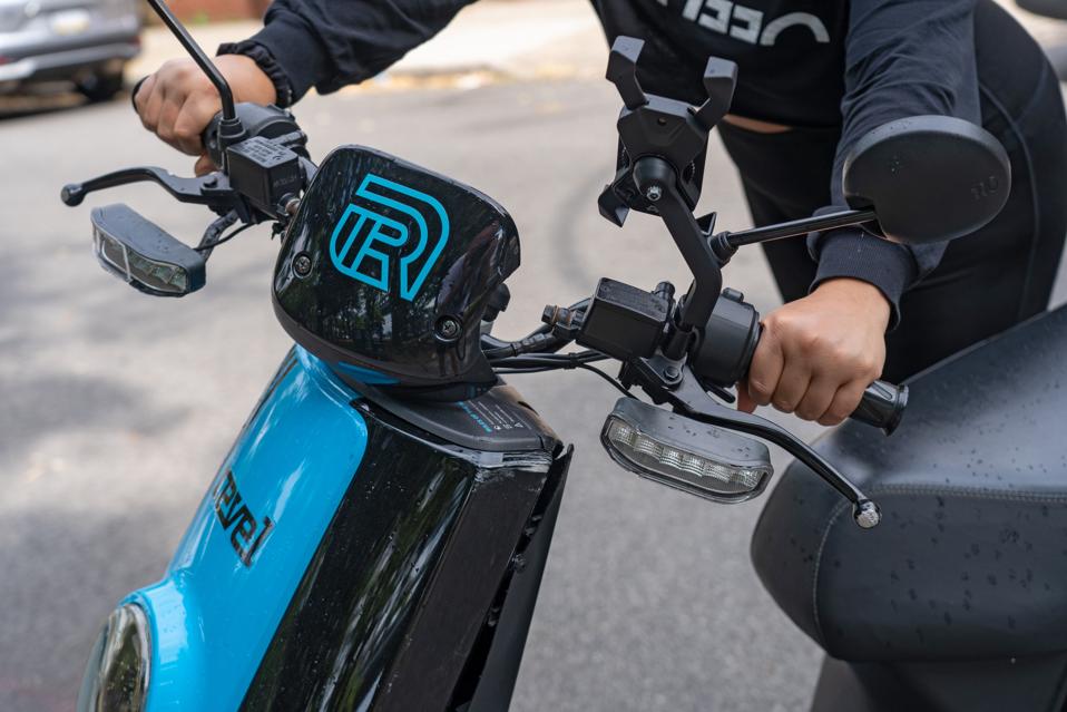 A woman holds handles of Revel moped on the street in the...