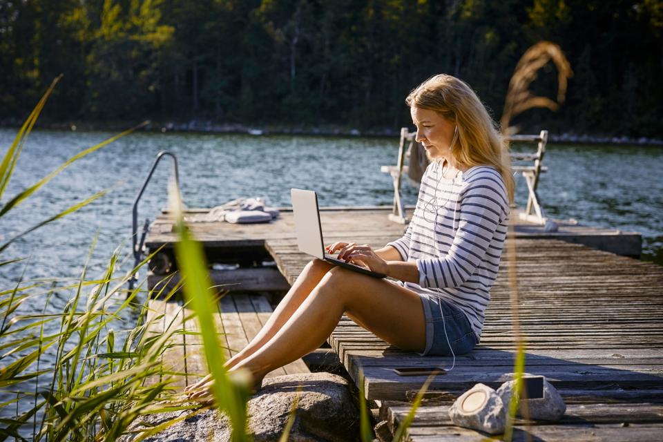 Woman with headphones using laptop while sitting on jetty against lake