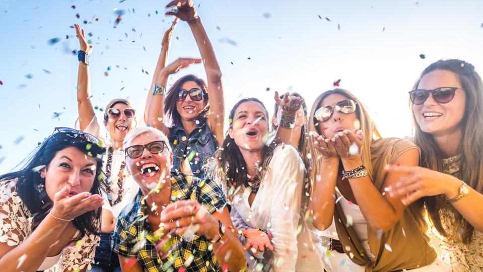Cheerful group of happy people mixed ages generations women having fun all together during celebration party or carnival - view of fiends blowing. coloured confetti and laugh a lot in friendship