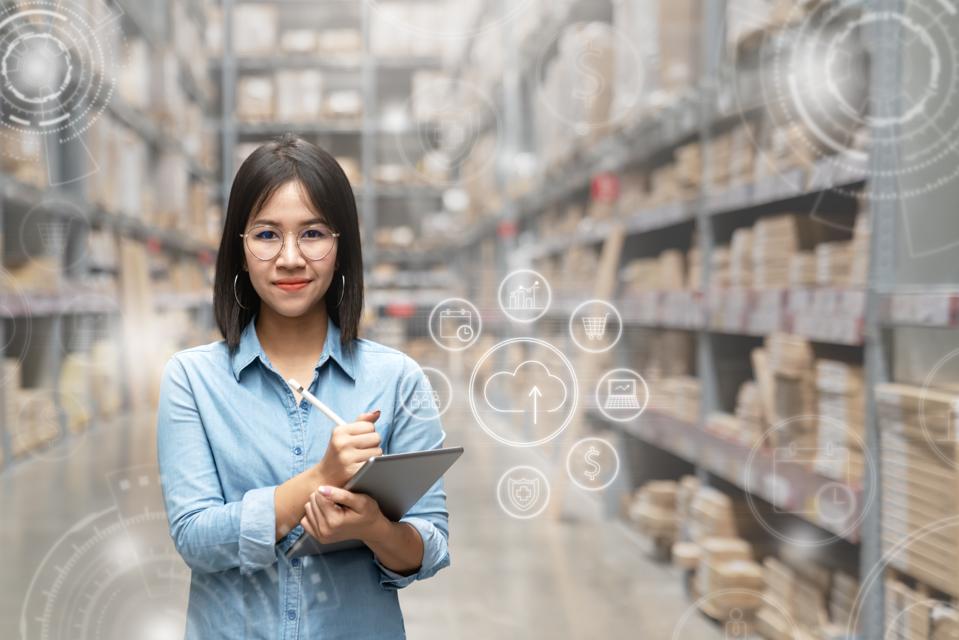 Portrait of happy young attractive asian entrepreneur woman looking at camera using smart tablet in warehouse with inventory management or industry digital era concept. Asian small business or sme.