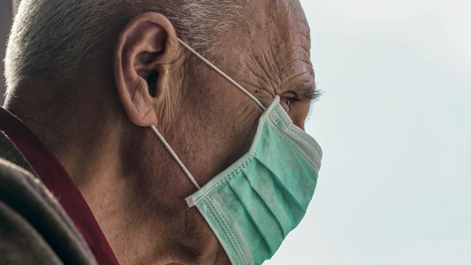 old man wearing medical face mask looking through window blue sky background