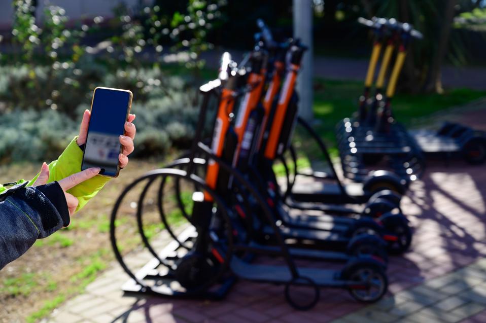 The faceless girl uses the mobile application to pay and unlock the rental electric scooter. Close-up female hand with a phone. Woman on eco-friendly transport to move around the city