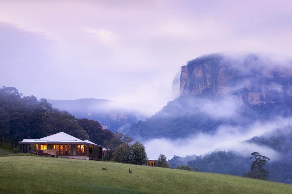 A luxe bungalow on a green lawn (with two rabbits), with a purple cloud-covered misty mountain in the background