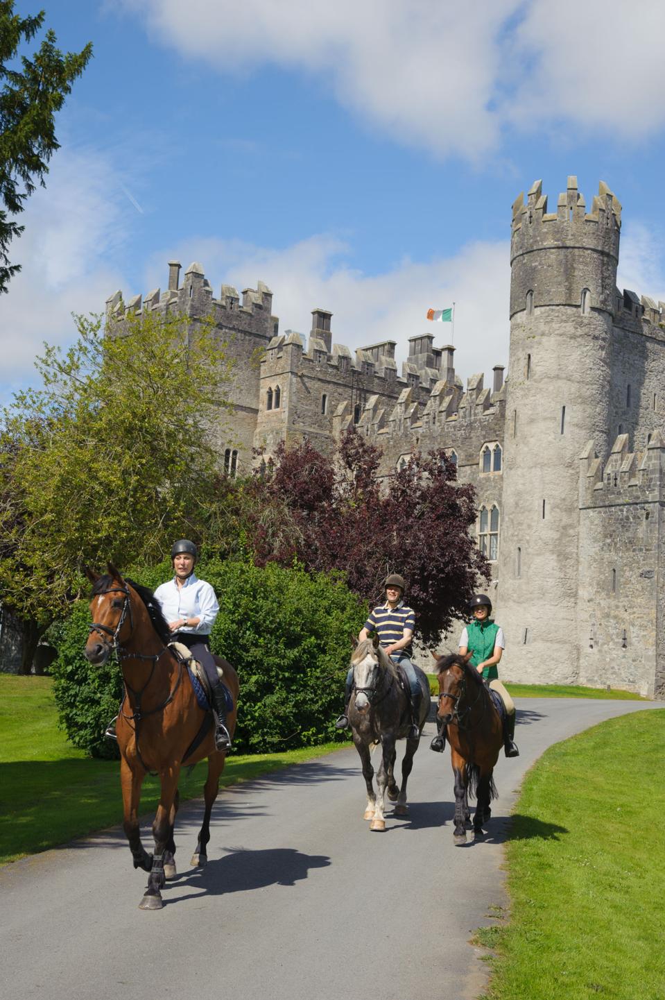 Three people on horseback on a path with  the stone walls and turrets of Kilkea Castle in the background