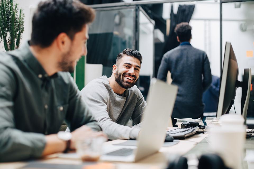 Colegas masculinos sonrientes mirándose mientras están sentados junto al escritorio en la oficina
