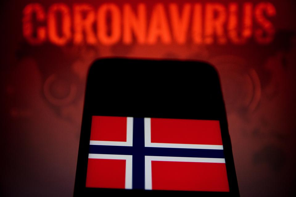 A Norway flag is displayed on a smartphone with the coronavirus warning in the background