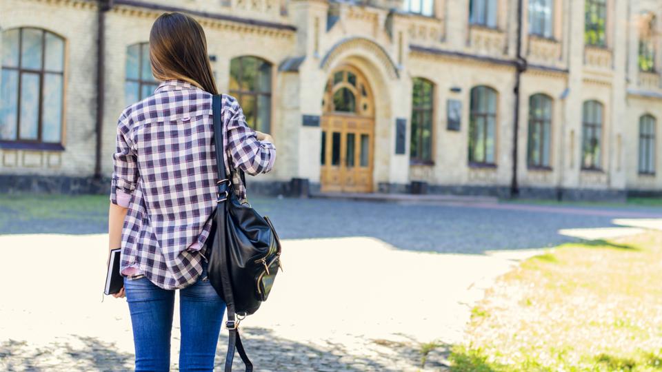 First day at university for young happy female student in casual clothes with backpack on her shoulders. View from back, she is looking at the entrance of the building