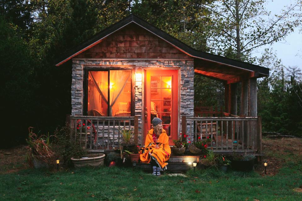 Woman wrapped in orange blanket sits outside cabin at dusk.