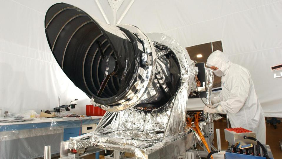 Color photo of an engineer in a white protective suit working on a large camera in a NASA lab.