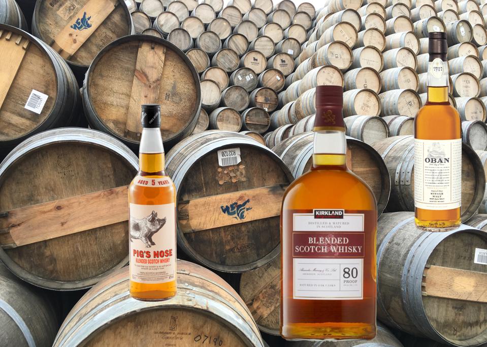 Three large bottles of scotch, Pig's Nose, Kirkland and Oban 14, sit atop scotch barrels