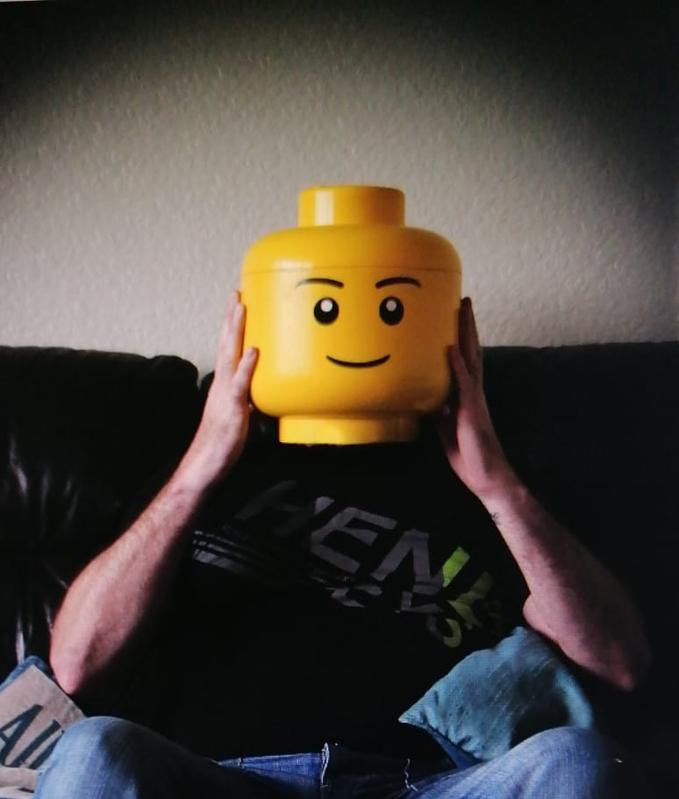 Man sits with lego character head in place of his own head represents masking of individuality