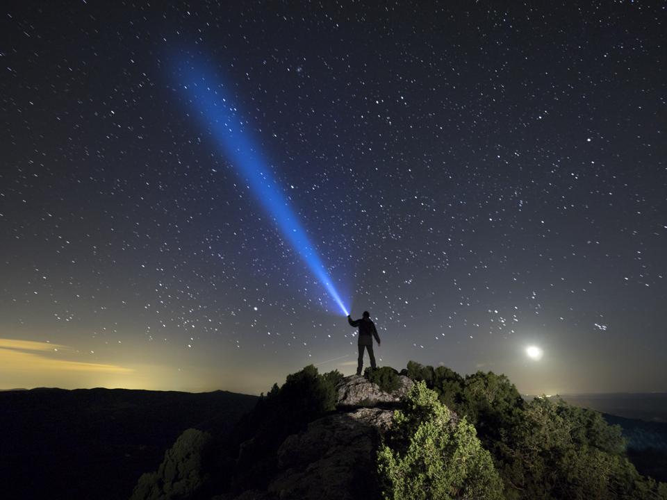 Silhouette of a man on the top of a mountain at night holding a beam of light up at the sky of stars