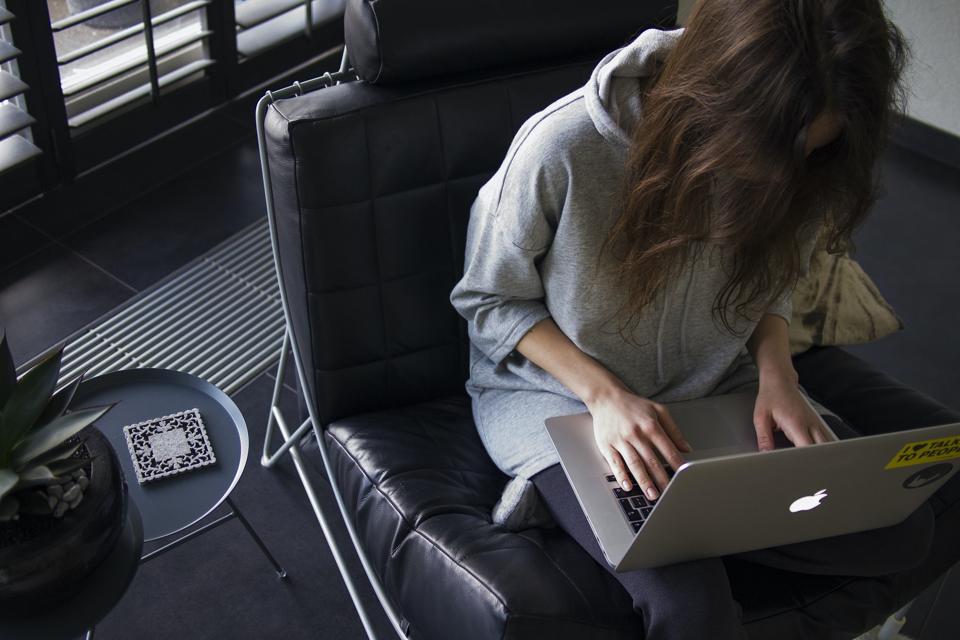 A woman sits on a black chair with her head down, typing on a computer.