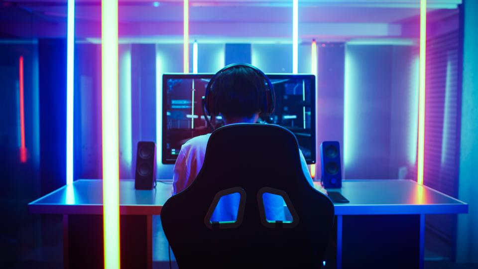 Back View Shot of the Beautiful Professional Gamer Girl Putting on Headset and Starts Playing Online Video Game on Her Personal Computer. Cute Casual Geek Girl. Room Lit by Neon Lamps in Retro Arcade Style.