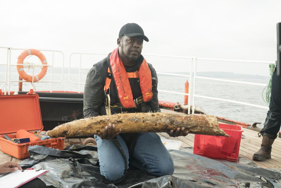 A coral-encrusted elephant tusk is held by a diver on a boat in the ocean.