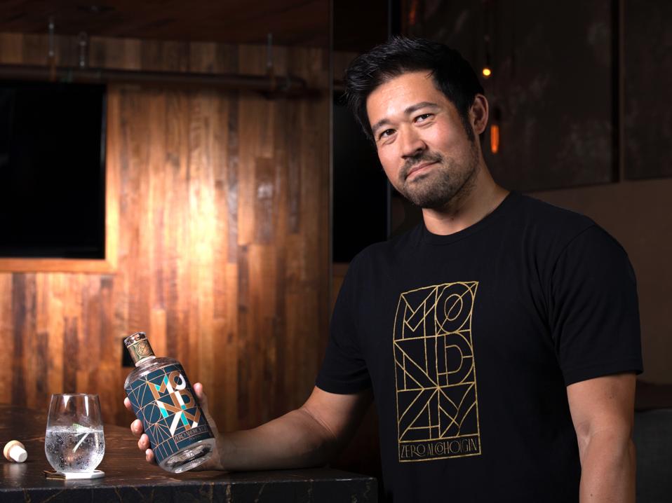 Boyd poses in a wood-paneled room holding a bottle of Monday Gin